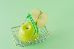 Ripe green apples on shopping toy basket on green background. Concept ofr grocery or market with copy space photo