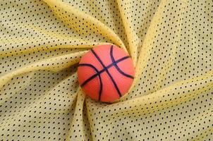 Small orange rubber basketball lies on a yellow sport jersey clothing fabric texture and background with many folds photo