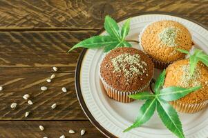 Healthy and tasty vegan and gluten free muffins topped with hemp seeds on a white plate on wooden table. Marijuana cupcake muffins with cannabis leaves photo