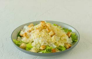 Traditional caesar salad in a plate on a white table with leaves, sauce, croutons and parmesan. photo