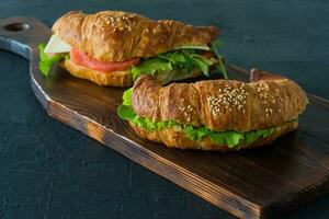 Croissant sandwiches with salted salmon on a desk, served with fresh salad leaves, arugula and vegetables over black background photo