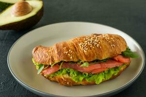 Croissant sandwich with salted salmon on a plate, served with fresh salad leaves, arugula and vegetables over black background photo