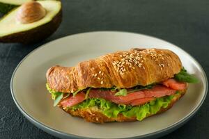 Sándwich de croissant con salmón salado en un plato, servido con hojas frescas de ensalada, rúcula y verduras sobre fondo negro foto