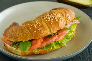 croissant sandwich with salmon, ricotta cheese and arugula on a plate on a dark background. Side view close up photo