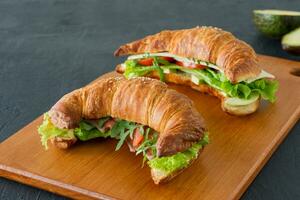 apetitosos sándwiches de croissant francés con salmón, aguacate, espinacas y lechuga, servidos en un escritorio de madera sobre un fondo gris. deliciosa comida para el desayuno. foto