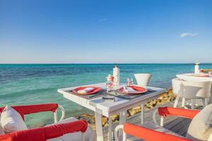 Luxury resort hotel over water outdoor restaurant on the beach, ocean and sky, seascape. Tropical island cafe, tables, food. Summer vacation or holiday, couple table setting with red white decoration