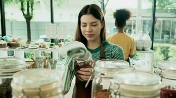 Young Caucasian female shopkeeper cleans glass jars of natural organic products in reusable containers at a refill store, zero waste, a plastic-free grocery shop, and an eco-friendly retail business.