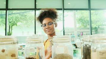 A young Black female shopkeeper cleans glass jars of natural organic products in reusable containers at a refill store, zero waste, a plastic-free grocery shop, and an eco-friendly retail business. video