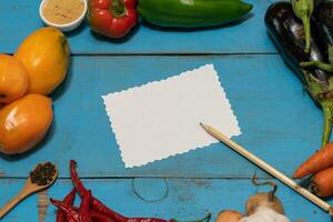 Vegetables are laid out around a sheet of paper and a pencil. Empty space for text. Vegetables, empty blank for recipe on a blue background. photo
