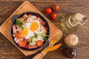 Homemade fried eggs with bacon and tomatoes in a frying pan on wooden background. Classic breakfast. Top view. photo