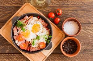 Homemade fried eggs with bacon and tomatoes in a frying pan on wooden background. Classic breakfast. Top view. photo