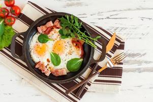 Overhead shot of breakfast or lunch with fried egg, spinach, arugula, tomatoes and bacon on black pan. photo