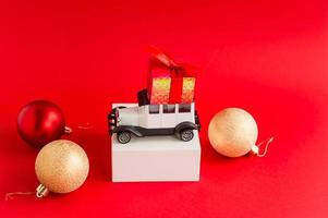 miniature model of a white retro car on a white pouill with a gift box on the roof. red New Year's background with Christmas tree colorful balls. photo