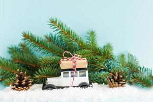 white toy retro car with a gift on the roof against the background of green spruce branches and cones in the winter snow. Christmas composition. photo