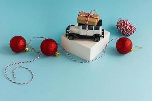 white toy retro car with a gift box on the roof of a blue New Year's background with red balls. photo