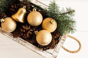 Christmas toys, gold balls and forest cones in a white wooden box with handles made of rope on a white wooden background. top view. close angle. photo