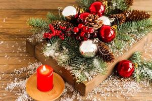 wooden box with red Christmas balls and branches, berries and cones on the village table with a Christmas candle. photo