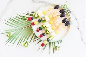 canapes of fresh fruit and berries on a plate. Marble background with palm tree branch. Festive food. photo