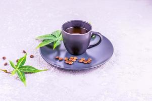 a dark cup with aromatic coffee and connabis leaves on a plate stands on a gray concrete background. The concept of morning coffee for energy. photo