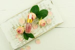 top view of the white wooden pedium with pink rose buds, green leaves and a cosmetic bottle of rose oil. natural facial skin care. photo