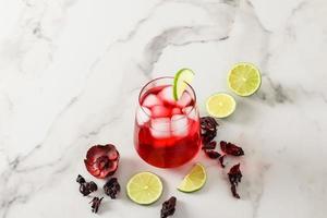 un vaso de bebida fría de hibisco con cubitos de hielo y gajos de lima sobre una mesa de mármol blanco. bebida tónica en el calor. foto