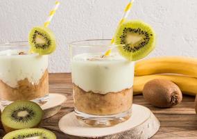 a glass of healthy homemade banana smoothie and kiwi with almonds on a wooden podium and a rustic table with fruit and a white wall. photo