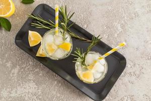 top view of two glasses of sparkling water with lemon and ice cubes on a ceramic tray and concrete table. fruit water is a concept of health. photo
