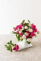 a beautiful bouquet of fresh garden roses in a decarative vase in the form of a watering can. concrete table against a white brick wall. photo