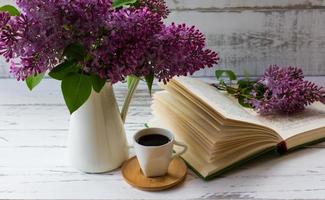 a bouquet of garden lilacs in a jug next to an open book and a cup coffee on a white background of wooden boards. photo