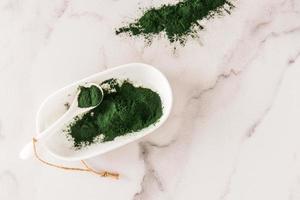 polvo de espirulina verde en un recipiente blanco ovalado con una cuchara sobre un fondo de mármol. el concepto de fortalecer el cuerpo. vista superior. foto