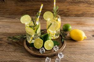 dos vasos de agua vitaminada con limón y lima, ramitas de romero en una bandeja redonda de madera y una mesa de pueblo. cubitos de hielo para enfriar.