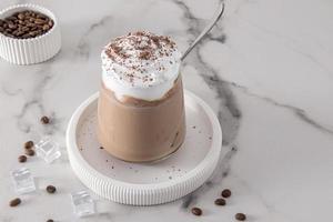 cold coffee with chocolate milk and ice, with whipped cream and cinnamon in a glass glass on a marble table. cold summer dessert. photo