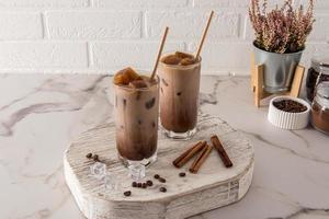 dos vasos altos con una bebida de chocolate o café frío con leche y cubitos de hielo se colocan en un podio de madera frente a una pared de ladrillo blanco. foto