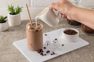 a woman's hand pours milk into a cold coffee in a modern glass in the form of a can. cold summer drink. chocolate pleasure.