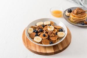 homemade breakfast. a large white bowl with mini pancakes with banana slices and blueberries on a wooden board and a white table. photo