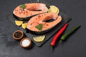 two raw salmon steaks with seasonings, lemon slices on a stone board and a black background. the concept of cooking a healthy meal rich in vitamins.