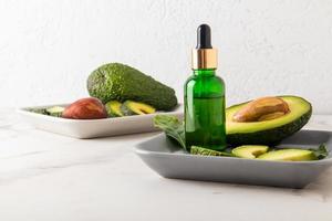 cosmetic oil with avocado extract in a bottle with a dropper on a ceramic rectangular plate with avocado fruit against a white wall. photo