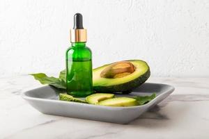 cosmetic avocado oil in a glass green bottle with a pipette on a ceramic gray tray with ripe avocado. natural cosmetics. photo