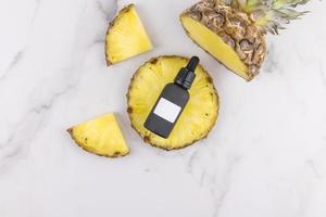 black matte cosmetic bottle with a natural organic product on a slice of pineapple and a marble background. Unbranded mock-up. photo
