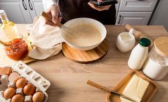 A woman's hand with a whisk beats the dough in a bowl. the other hand holds the phone with the recipe for further cooking. photo