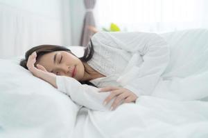 A beautiful woman lays her hands on a soft pillow. A young woman wearing white pajamas lies under a warm blanket. photo