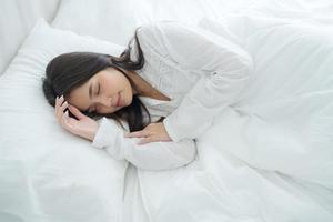 una mujer hermosa pone sus manos sobre una almohada suave. una mujer joven con pijama blanco yace debajo de una manta cálida. foto