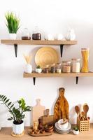 front vertical view of open kitchen wooden shelves and parts of the countertop with various kitchen utensils, storage jars, plants in pots. photo