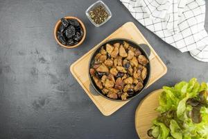 stew with prunes in a frying pan is served on a wooden board with fresh lettuce greens. top view. black background. gourmet food. photo