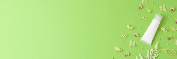 White tube of cosmetic cream with flowers on green background. Flat lay, copy space photo