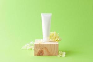 White tube of cosmetic cream with wood podium and flowers on green background. Close up, copy space photo