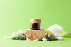 Jar of cosmetic cream with wood podium and flowers, moss, stone on green background. Close up, copy space photo