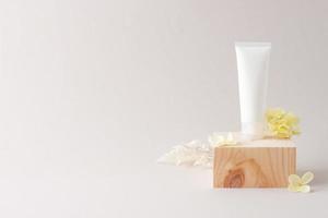 White tube of cosmetic cream with wood podium and flowers on grey background. Close up, copy space photo