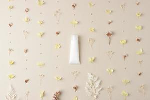 White tube of cosmetic cream with flowers on pastel beige background. Flat lay photo