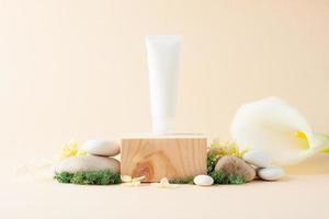 White tube of cosmetic cream with wood podium and flowers, moss, stone on pastel beige background. Close up, copy space photo